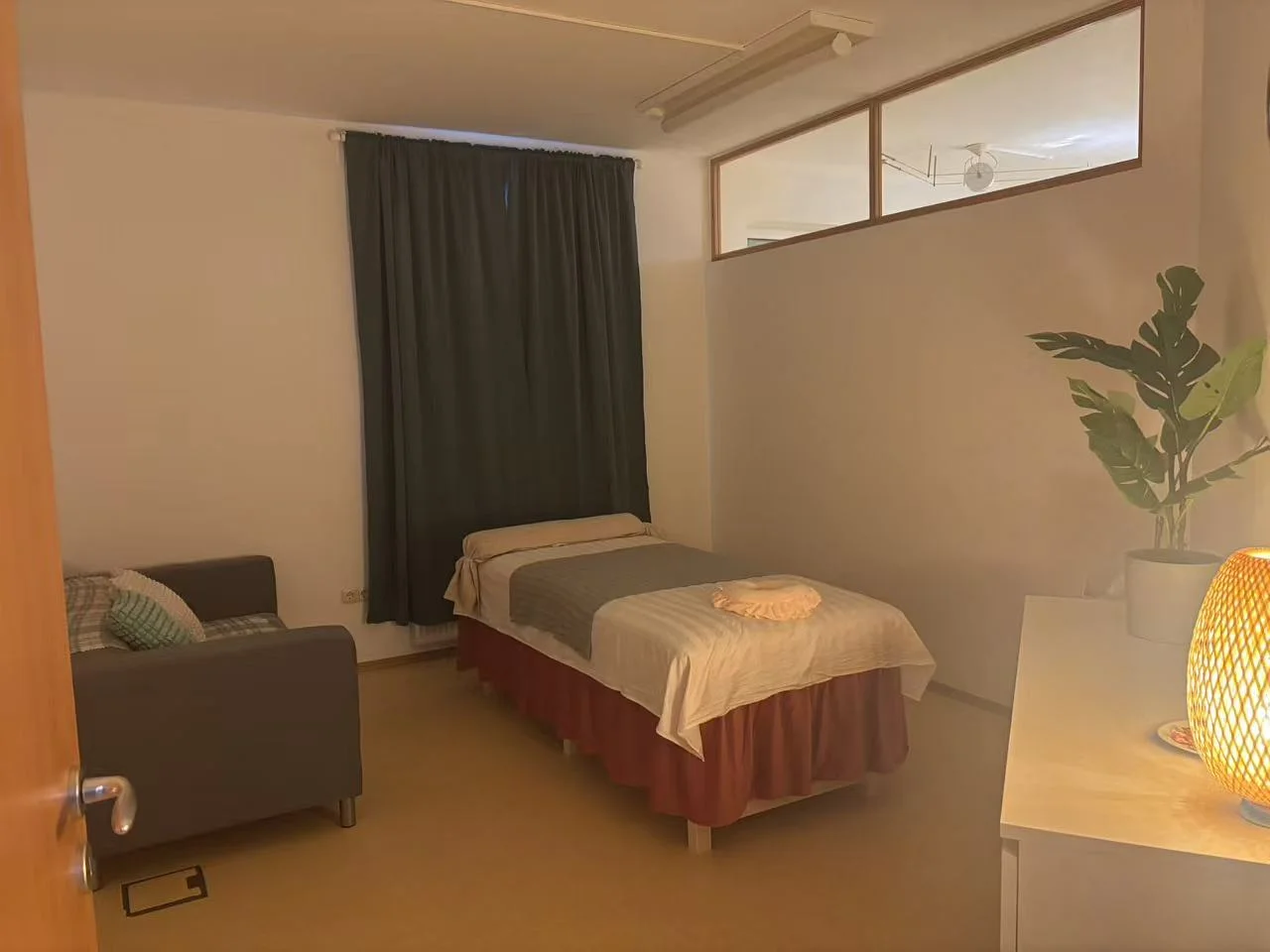 Massage treatment room with a white-covered massage bed, light gray runner, and decorative orange circular pillow. A small lamp provides warm ambient lighting.