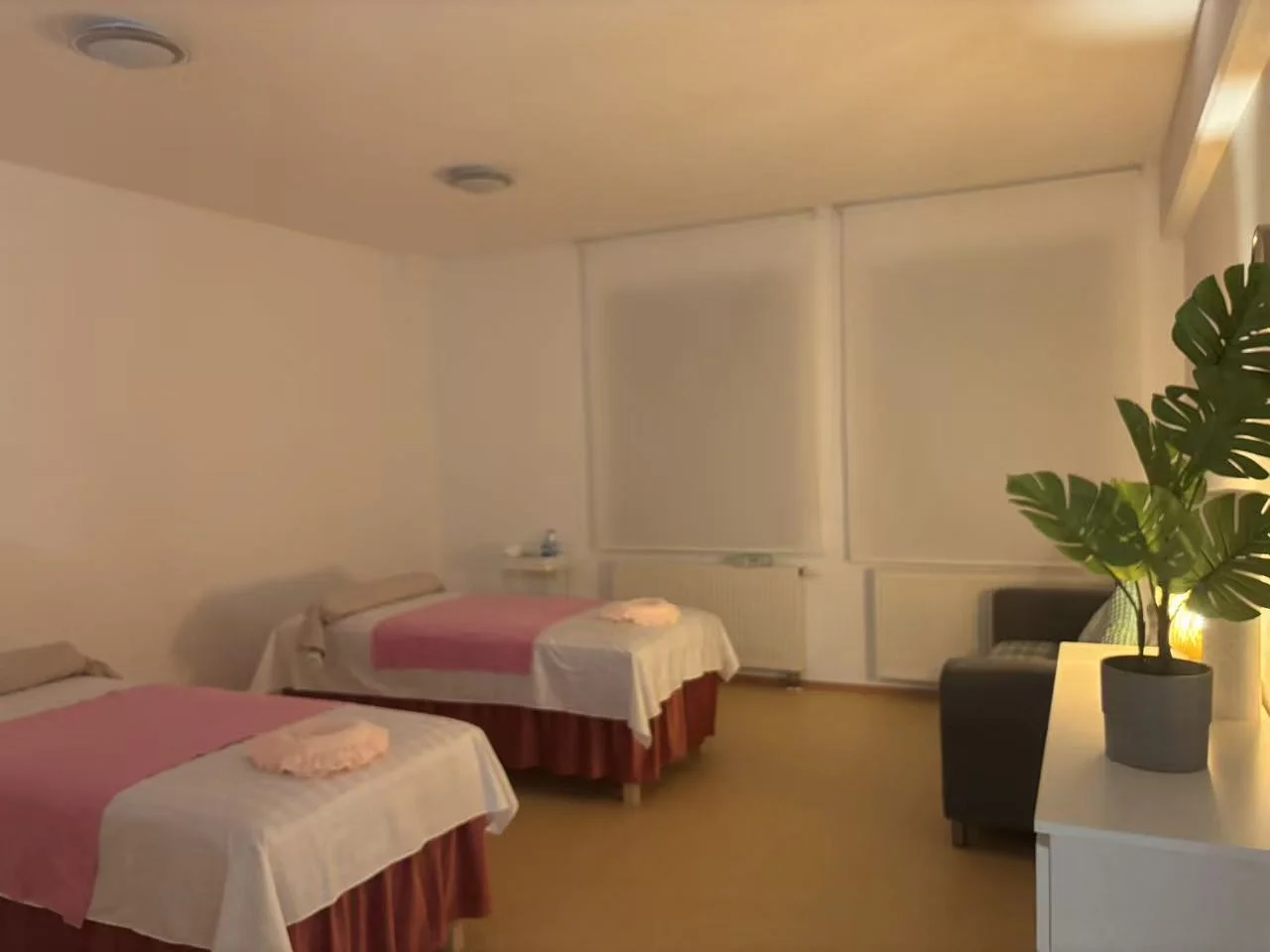 Treatment room with massage bed, decorative pillows, small storage cabinet, wall-mounted clock, and coat hooks. Door leads to hallway.